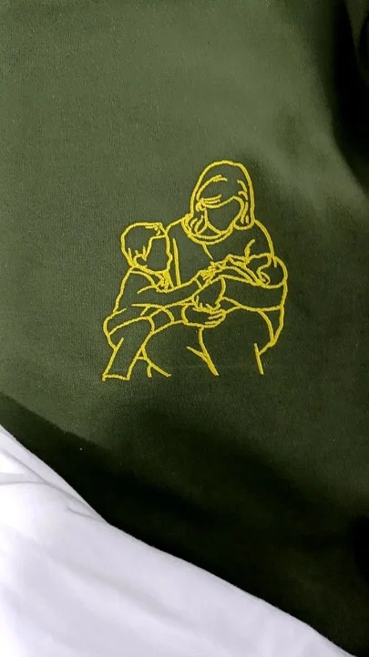 Kundin zeigt olivgrünes T-Shirt mit gelber Stickerei von Mutter und zwei Kindern